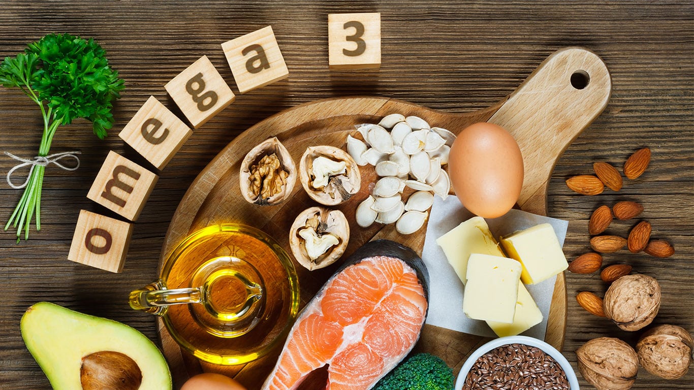 oomega 3 spelled out with wooden blocks next to an array of foods including fish, nuts, seeds, eggs, butter, avocado, and parsley